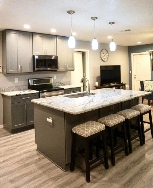 Modern kitchen with gray cabinets, stainless steel appliances, a large marble island with a sink, three cushioned bar stools, pendant lights overhead, and a living area with a TV and wall clock in the background.