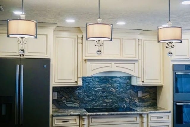 A modern kitchen with cream-colored cabinets, a blue and gray marble backsplash and countertop, three pendant lights, a black refrigerator, and a double wall oven.