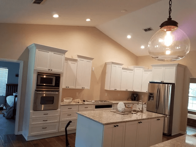 Modern kitchen with white cabinets, stainless steel appliances, a kitchen island with a marble countertop, and a glass pendant light. There are dishes on the island and a bedroom visible through an open doorway.