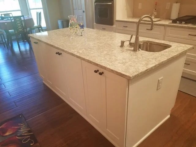 A kitchen island with a light-colored granite countertop, white cabinets, a built-in sink, and a faucet. The island sits on a wooden floor and is adjacent to a kitchen and dining area.