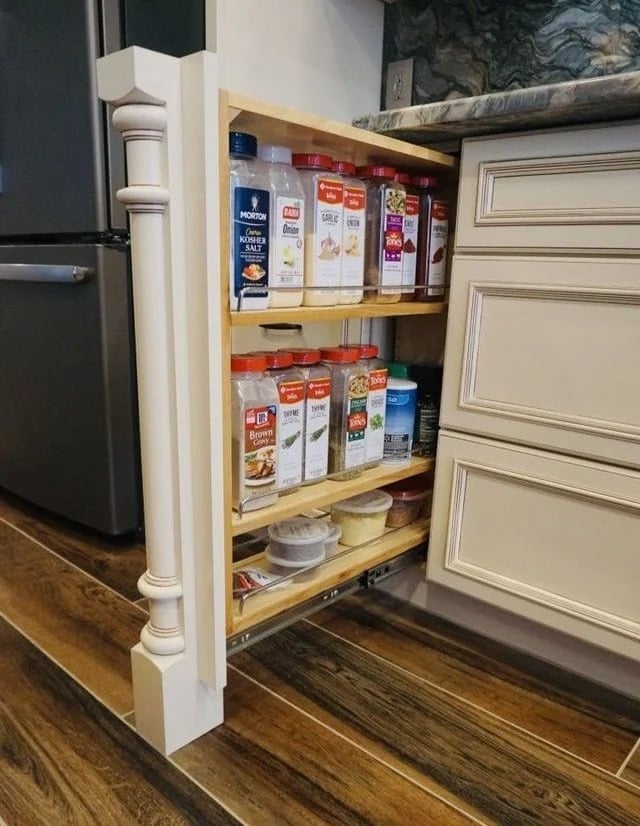 A narrow pull-out kitchen cabinet with three shelves holds various spice containers and jars, neatly organized beside a fridge and counter on a wood floor.