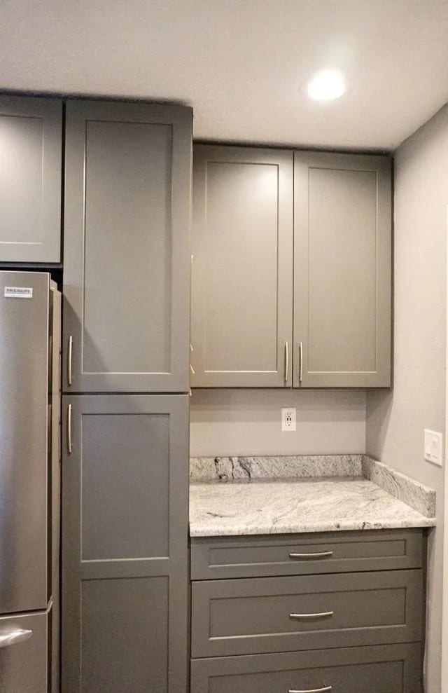 A modern kitchen corner with gray cabinets, silver handles, a gray and white marble countertop, and a stainless steel refrigerator partially visible on the left. There is an overhead light and electrical outlets on the wall.