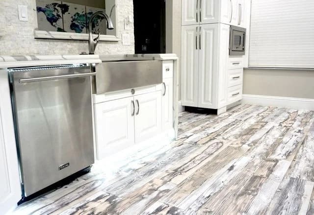 Modern kitchen with white cabinets, a stainless steel dishwasher, farmhouse sink, and rustic whitewashed wood flooring. The kitchen is bright and features a built-in microwave and large windows with blinds.