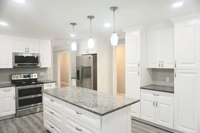 A modern kitchen with white cabinets, granite countertops, stainless steel appliances, a large island, and pendant lights hanging from the ceiling. The floor is gray wood-patterned, and the space is brightly lit.