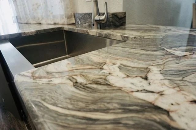 A close-up view of a kitchen countertop with a striking, swirling gray, white, and black marble pattern, next to a stainless steel sink and a modern faucet.
