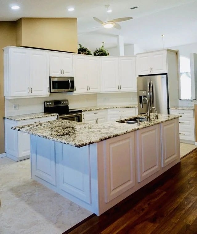 Modern kitchen with white cabinets, granite countertops, a large island with a sink, stainless steel appliances, and a mix of tile and wood flooring. Ceiling fan and greenery displayed above the cabinets.
