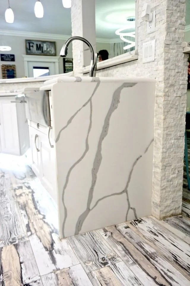A modern kitchen features a white marble sink with gray veining, matching a white cabinet and rustic, weathered wood flooring. The background includes white walls, pendant lights, and decorative wall art.