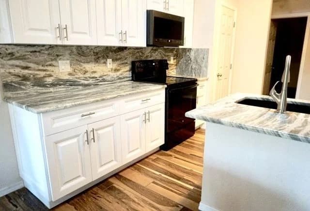 Modern kitchen with white cabinets, black appliances, grey and white marble countertops and backsplash, and a wood floor. A sink with a sleek faucet is on the kitchen island in the foreground.