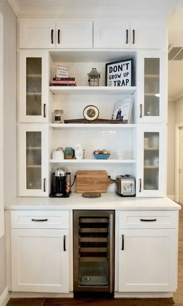 A white built-in kitchen cabinet features glass-paneled cabinets, open shelves with books and decor, a coffee maker, a toaster, a wine fridge, and a sign that reads "DON'T GROW UP IT'S A TRAP.