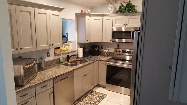 Modern kitchen with light gray cabinets, granite countertops, stainless steel appliances including a stove, microwave, and dishwasher, a sink, small appliances, and decorative items on the cabinets. White tile floor.