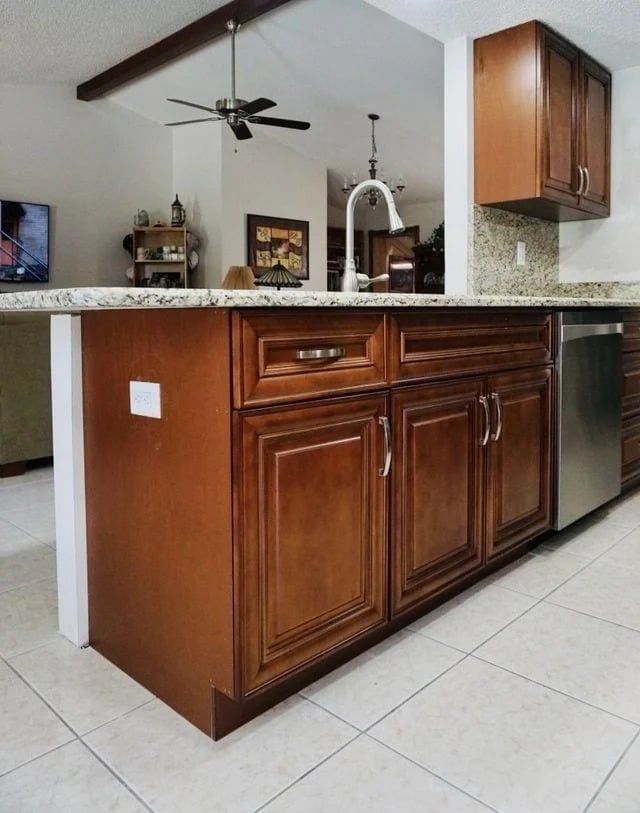 A kitchen island with dark wood cabinets and silver handles, granite countertop, dishwasher, and sink. The floor is tiled, and the background shows part of a living area with ceiling fan and wall art.