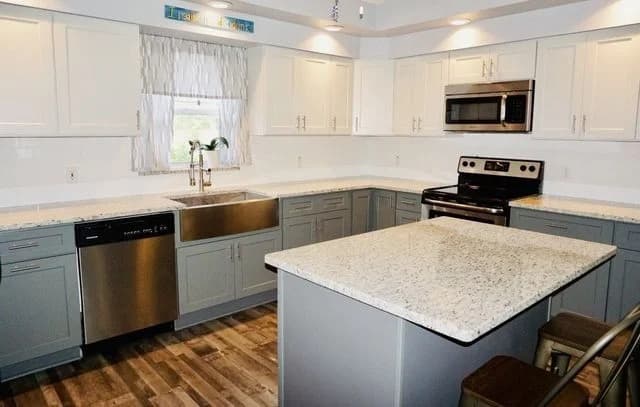Modern kitchen with white and gray cabinets, stainless steel appliances, a farmhouse sink, center island with granite countertop, and wood-look flooring. A window with sheer curtains is above the sink.