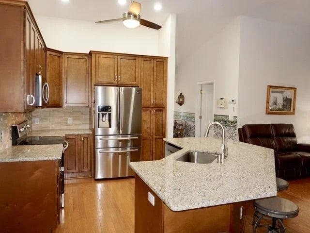Modern kitchen with wooden cabinets, stainless steel appliances, a double-door refrigerator, granite countertops, and an island with a sink and barstools. Part of a living area with brown leather seating is visible.