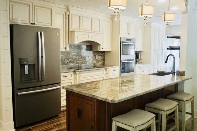 A modern kitchen with cream cabinets, stainless steel appliances, a marble island with a sink, and three upholstered stools. Pendant lights hang above the island, and the backsplash features a gray stone design.