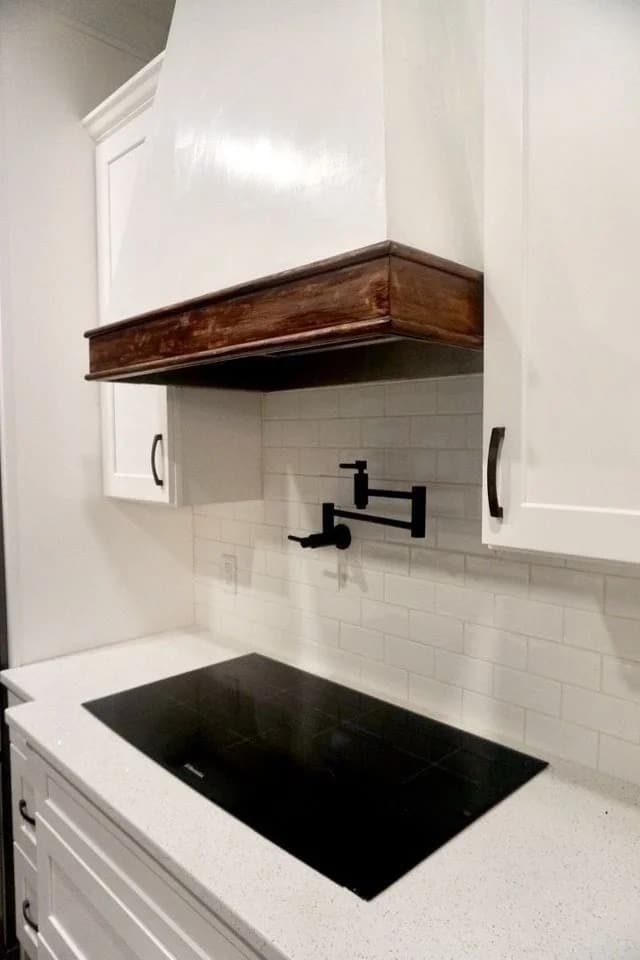 Modern kitchen with a white tile backsplash, white cabinets, a black stove top, a wall-mounted black pot filler faucet, and a white vent hood with a dark wooden trim.