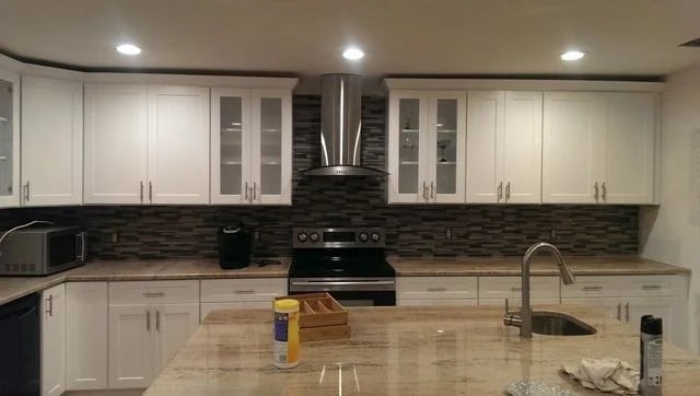 Modern kitchen with white cabinets, stainless steel oven and range hood, dark tile backsplash, and a large island with a sink. Cleaning supplies and a wooden organizer are on the countertop.