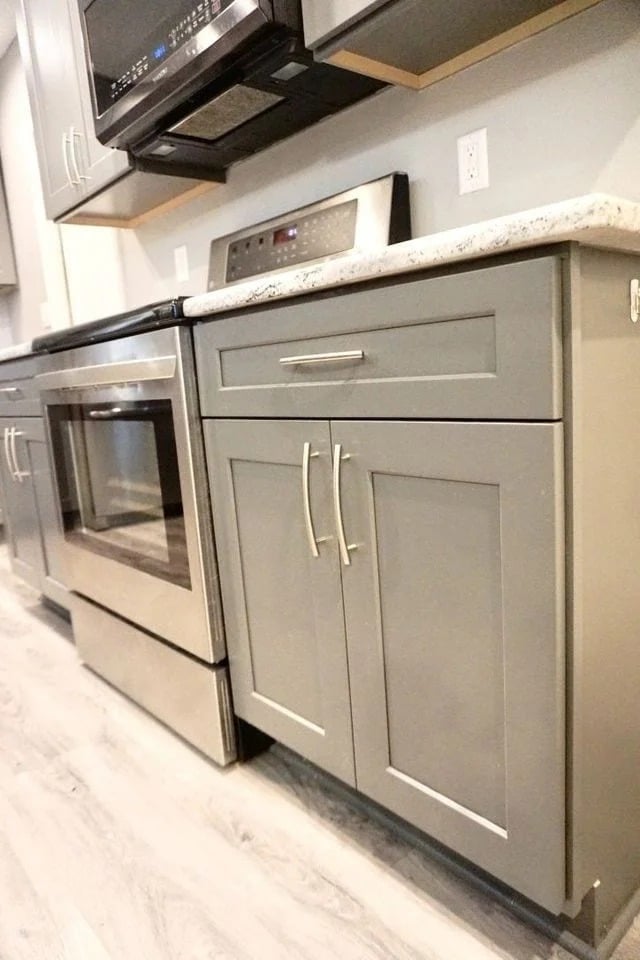 A modern kitchen with light gray cabinets, stainless steel appliances, a granite countertop, and a drawer and cabinet below the counter. The floor is light-colored wood or laminate.