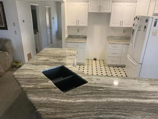 A kitchen with white cabinets, marble-patterned countertops, a black sink, and a white refrigerator. The floor has a white and black tile pattern, and the countertop has unique, flowing gray and beige streaks.