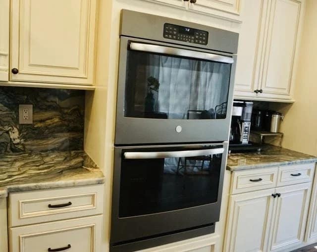 A modern built-in double wall oven is installed between cream-colored cabinets and drawers with marble countertops and backsplash. A coffee maker and other kitchen items are visible on the right countertop.