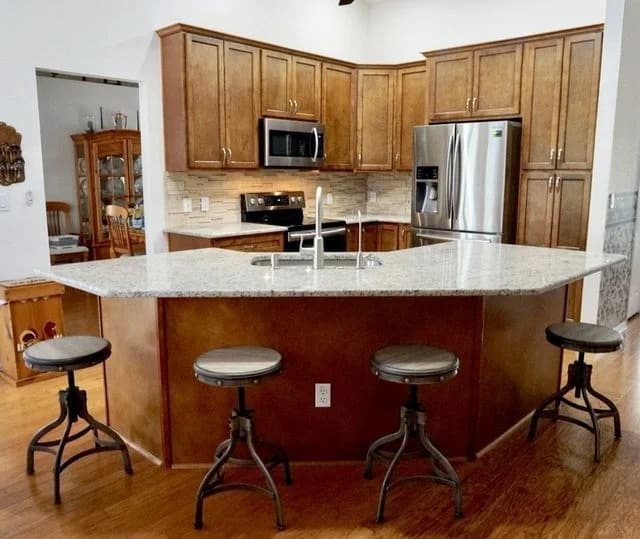 A modern kitchen with wooden cabinets, granite countertops, stainless steel appliances, and an island with a sink. Four metal barstools with wooden seats surround the island on a wooden floor.
