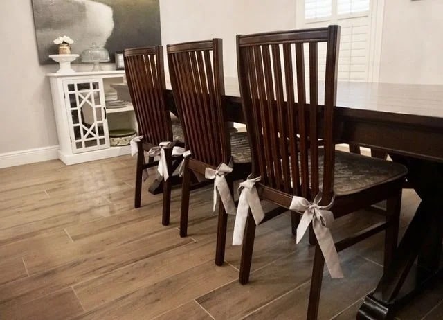 Three wooden dining chairs with vertical slats and patterned cushions are tied with decorative gray bows at the back. They are placed around a dark wooden dining table on a light wood floor in a modern dining room.