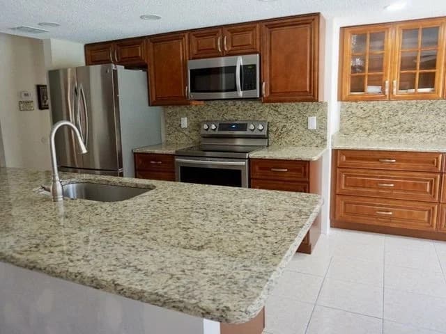 Modern kitchen with stainless steel appliances, wooden cabinets, granite countertops and backsplash, a built-in microwave, electric stove, glass-front cabinets, and a sink on a center island. White tile floor.