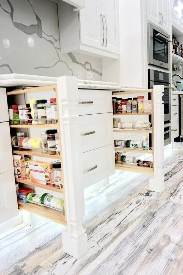 A modern kitchen features two white pull-out spice racks built into an island, displaying organized jars and cans. The floor has a light wood pattern, and sleek white cabinets and appliances are visible in the background.