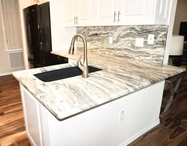 A modern kitchen counter with a marble-patterned surface, integrated black sink, and silver faucet. The backsplash matches the countertop, and white cabinets are above. The floor is wooden, and appliances are black.