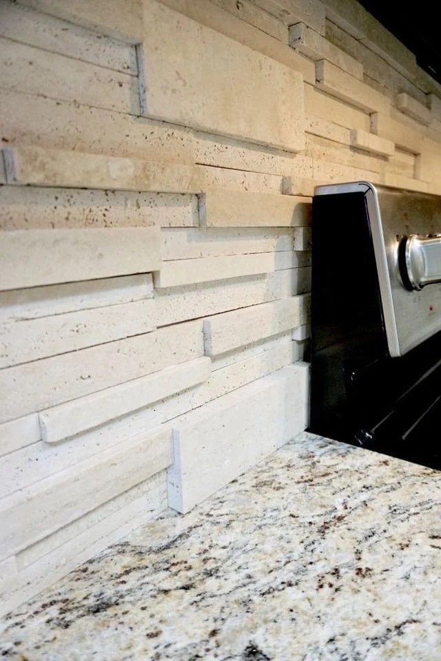 A kitchen countertop with a beige, textured 3D tile backsplash featuring staggered rectangular tiles, next to a stainless steel stove. The countertop surface has a speckled, granite-like pattern.