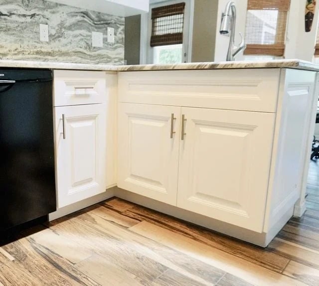 White kitchen cabinets with silver handles are set against a marbled countertop. A black dishwasher is installed beside the cabinets. The floor is wood with a natural finish, and the room is bright with natural light.