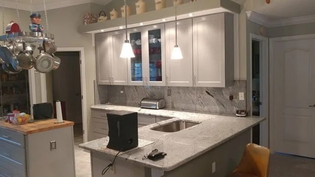 A modern kitchen with gray cabinets, marble countertops, a hanging pot rack, and pendant lights over the island. A small TV, toaster, and other kitchen items are on the counter. The walls are light-colored.