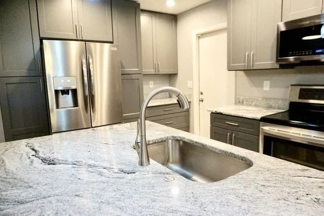 Modern kitchen with gray cabinets, stainless steel appliances, a built-in refrigerator, and a marble countertop island featuring an under-mount sink and sleek faucet. The space is brightly lit with recessed lighting.