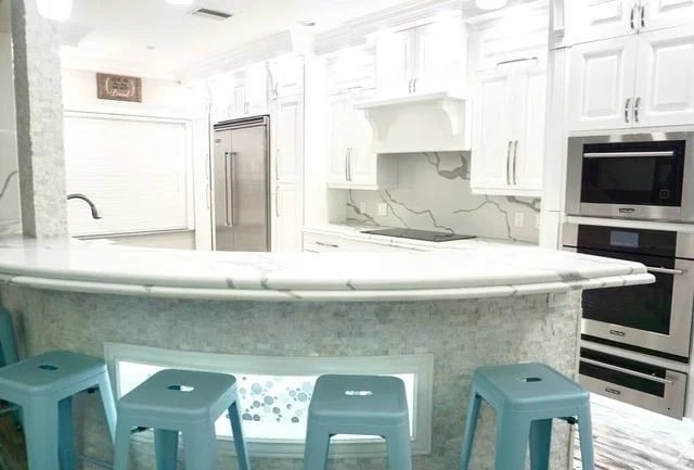 A modern white kitchen with marble countertops, stainless steel appliances, a double oven, and a curved breakfast bar with blue stools. The backsplash features a gray and white marble pattern.