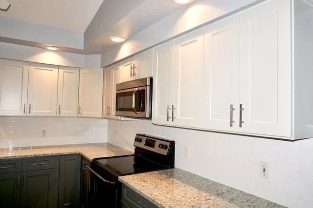 A modern kitchen with white upper cabinets, dark lower cabinets, granite countertops, a stainless steel stove, a built-in microwave, and white subway tile backsplash under recessed ceiling lights.