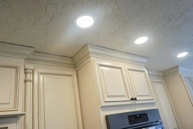 Cream-colored kitchen cabinets do not reach the textured ceiling, leaving a noticeable gap. Recessed ceiling lights are turned on, and a stainless steel oven is partially visible below the cabinets.
