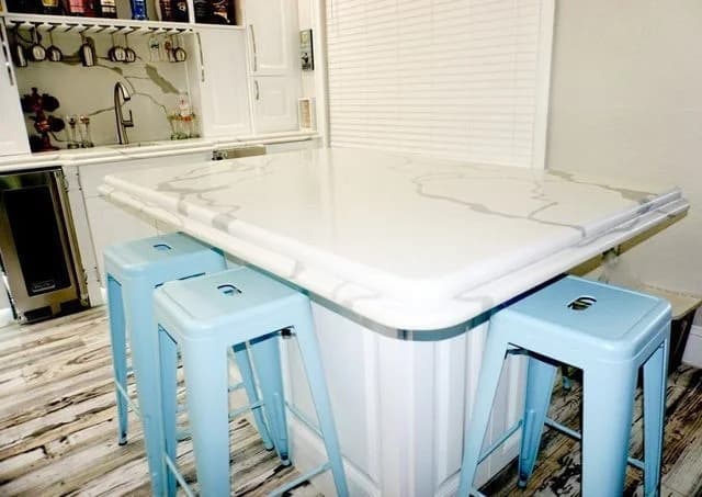 A modern kitchen features a white marble island with gray veining, surrounded by four light blue metal stools on a wood-patterned floor. The background includes a sink, shelves, and closed white blinds.