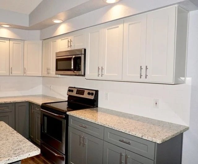 Modern kitchen with white upper cabinets, gray lower cabinets, granite countertops, stainless steel appliances (oven and microwave), under-cabinet lighting, and white subway tile backsplash.