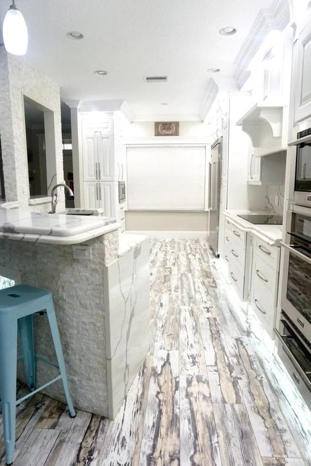 A modern kitchen with white cabinets, marble countertops, textured white backsplash, a farmhouse sink, and wood-look flooring. A blue metal stool sits by the counter, and there are built-in ovens on the right.