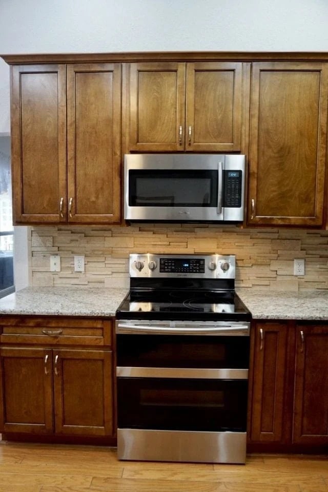 A kitchen with wooden cabinets, a stainless steel microwave above a matching electric stove, granite countertops, and a beige tile backsplash. The floor is wood and there are electrical outlets on the wall.