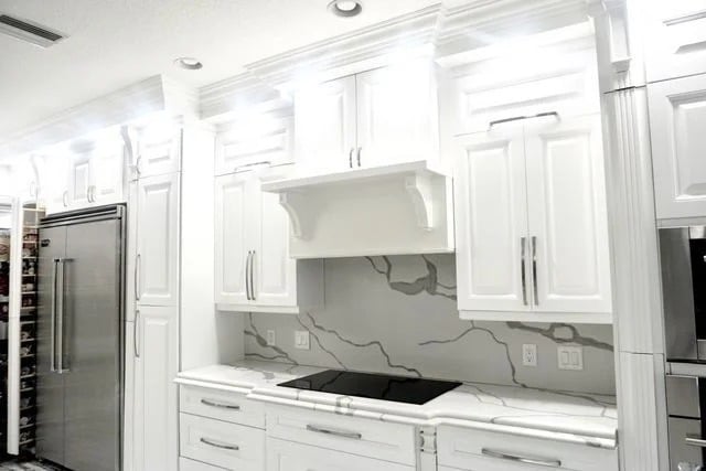 Bright, modern kitchen with white cabinets, stainless steel refrigerator, marble-patterned backsplash and countertops, and an electric cooktop beneath a vent hood. Recessed ceiling lights illuminate the space.