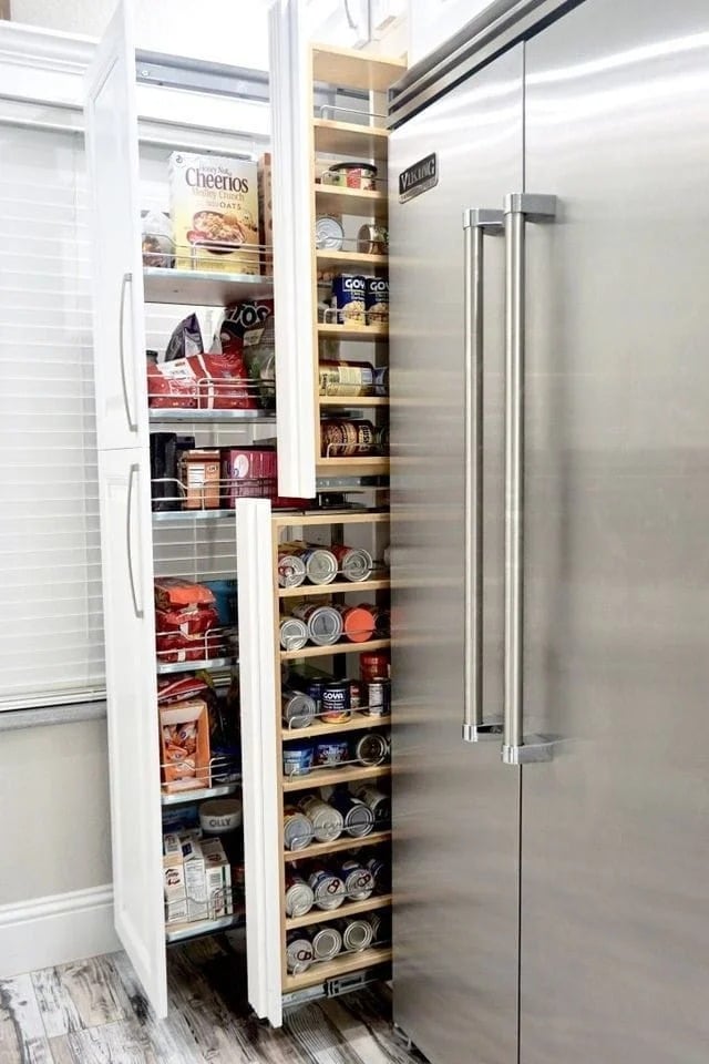 A tall, narrow pull-out pantry next to a stainless steel refrigerator is filled with cereal boxes, canned goods, and snacks, efficiently organized on several shelves.