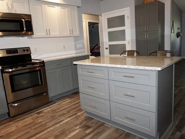 Modern kitchen with stainless steel appliances, gray cabinets, a large kitchen island with drawers, white countertops, and wood-look flooring. White upper cabinets and a door with frosted glass panels are visible in the background.