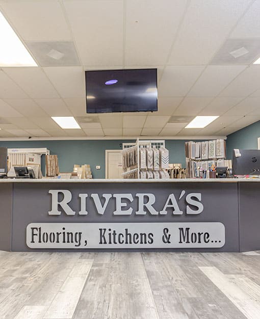 A showroom reception desk with a large sign reading "RIVERA'S Flooring, Kitchens & More..." beneath a mounted TV, with flooring samples displayed behind the counter.