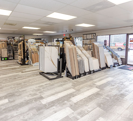 A flooring showroom with displays of various tile and flooring samples arranged in rows on stands, under bright ceiling lights on a wood-look floor, with large windows letting in natural light.