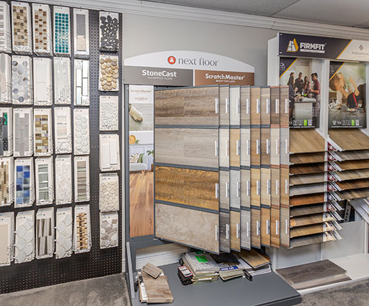Display racks in a flooring store showcase various samples of tile, stone, and wood flooring options. A central stand presents large wood-look planks while adjacent displays feature more samples and brochures.
